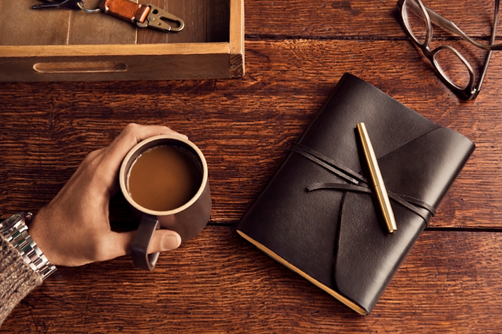 Una mano sostiene un pocillo de café sobre un escritorio de madera, cerca de unas gafas y un diario de viaje forrado en cuero oscuro